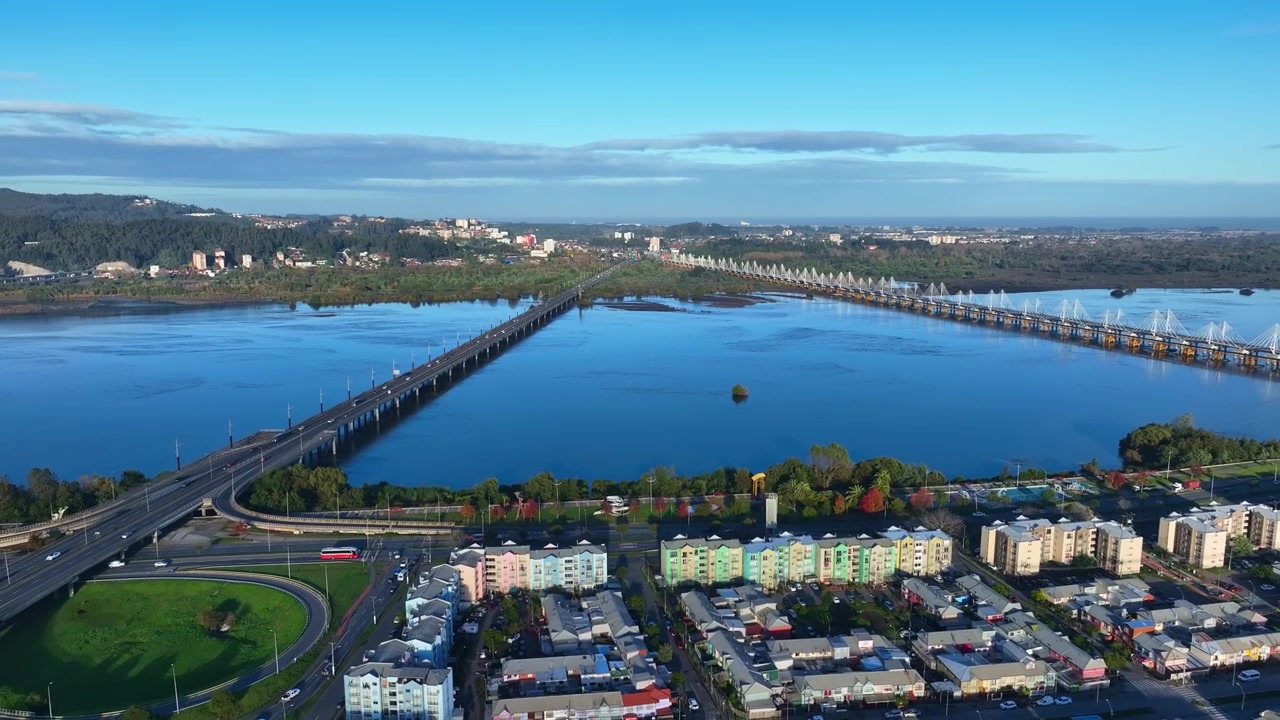 Ripresa aerea che rivela due ponti che attraversano l'ampio fiume Bio Bio con colorati condomini lungo il fiume, svincolo autostradale e rive boscose sotto un cielo azzurro e limpido. Concepcion, Cile.