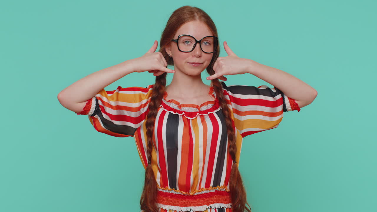 niña pelirroja mirando a la cámara haciendo un gesto telefónico como dice hola me llamas, número de contacto