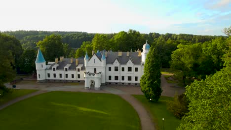 aerial drone footage orbiting around a gorgeous and majestic white colored alatskivi castle that has large towers during sunset at summer time. green forest and treetops around the historic building