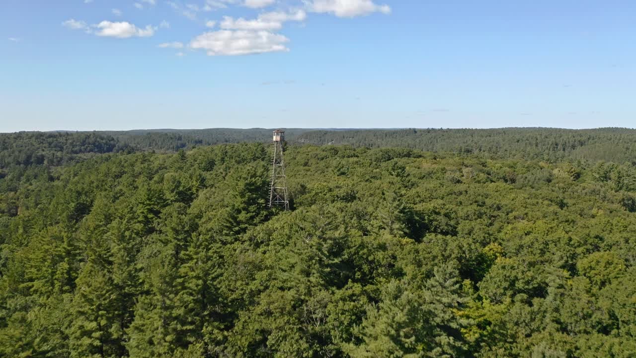 Drone flying over treetops toward lookout tower on blue sky day