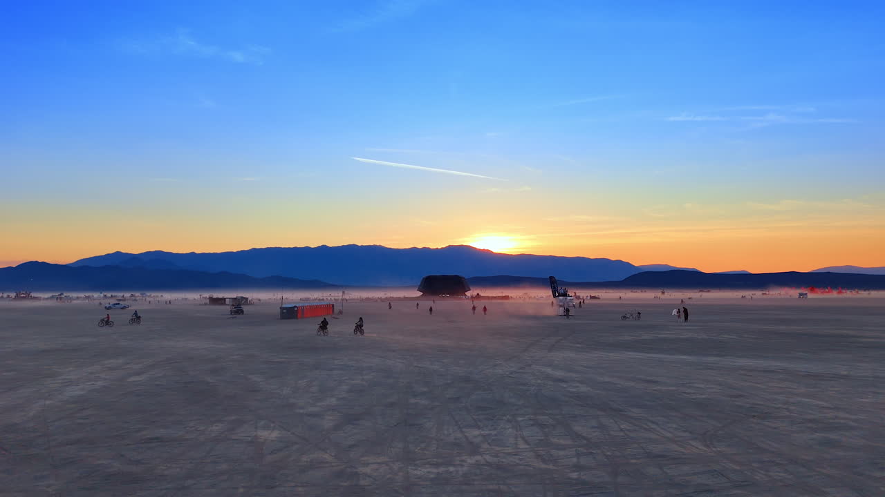 Sunset haze over wide desert festival zone. A fading sun casts soft colors over mountains and distant gatherings scattered across the open playa