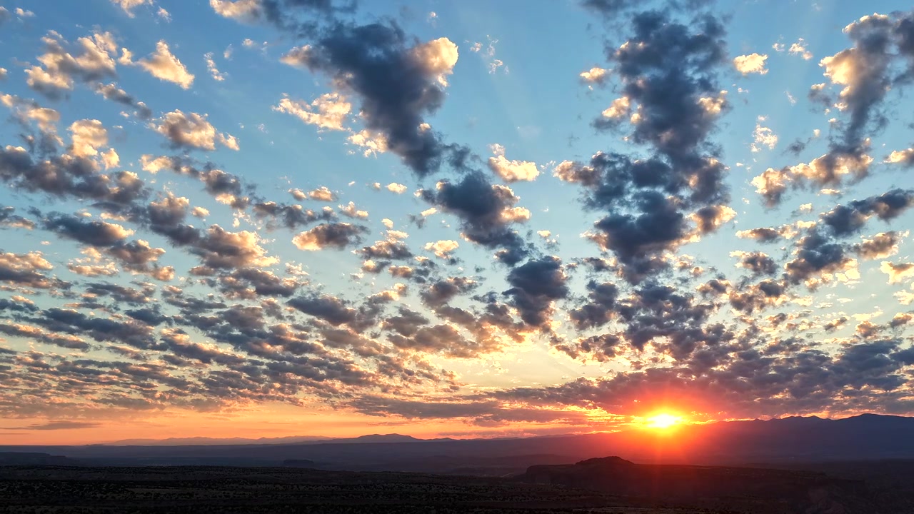 Timelapse 4K da drone di un'alba vibrante che irrompe su un paesaggio aspro di montagne e canyon. Distinte file di nuvole si muovono ritmicamente mentre raggi solari radiosi si proiettano verso l'alto nel cielo blu dell'alba.