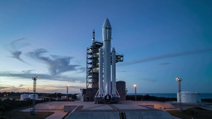 Standing three-core rocket awaiting launch on coastal pad as dusk falls, gantry and lights glowing