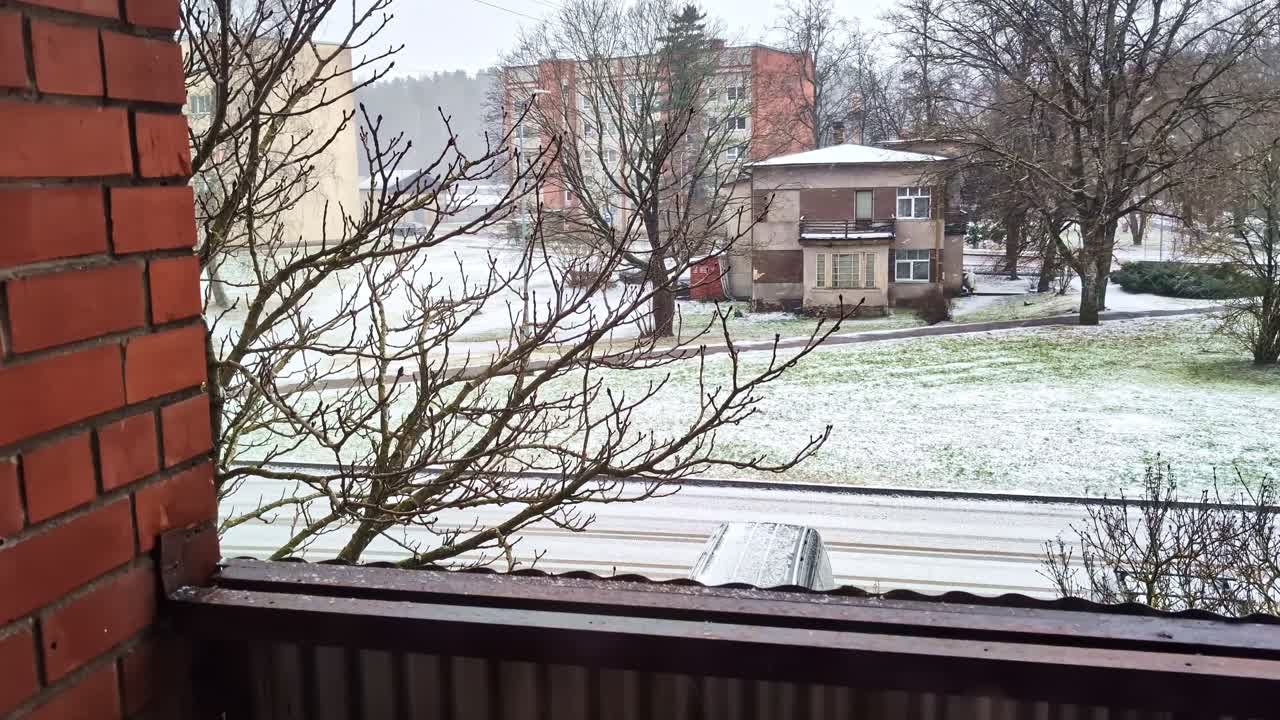 Urban landscape view from balcony with bare tree branches and snow on ground during snowfall