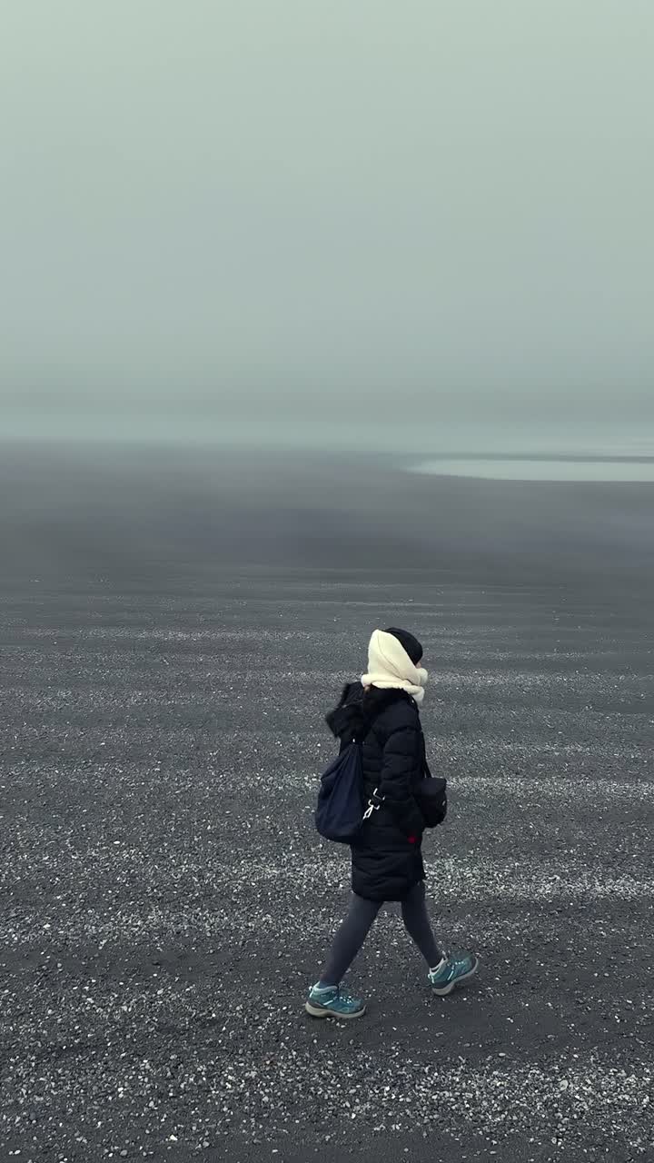 Donna con giacca invernale che cammina su una spiaggia di sabbia nera vulcanica e nebbiosa a Stokksnes.