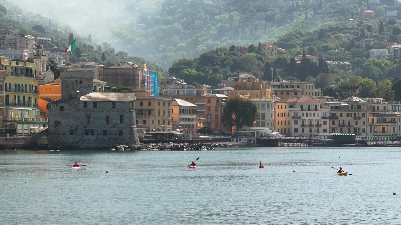 Ampia inquadratura tranquilla di kayakisti che remano vicino al Castello di Rapallo, colorati edifici liguri e rigogliose colline sotto una morbida luce naturale. Vivace paesaggio marino urbano della Riviera Ligure