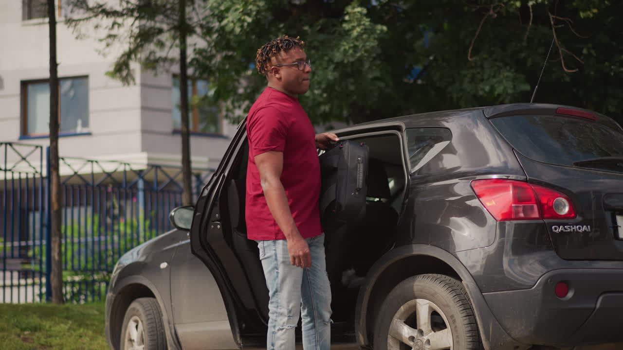 Uomo scarica una borsa dal bagagliaio dell'auto su un marciapiede urbano fuori da un complesso di appartamenti, musicista maschio rimuove la borsa dell'attrezzatura, controlla i cavi, chiude il bagagliaio, cammina verso la strada con passo deciso, luce calda.