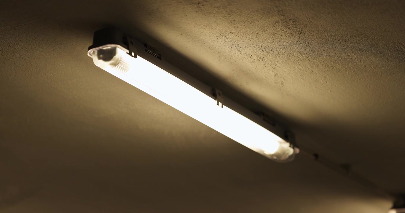 Turning on a fluorescent ceiling lamp from darkness to full brightness in a bare garage interior space.