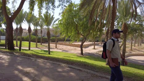 a man is walking down a path in a park. he is wearing a backpack and a hat.