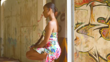 una chica sexy con un vestido se pone en cuclillas sobre una viga en un almacén abandonado con graffiti en la pared de fondo