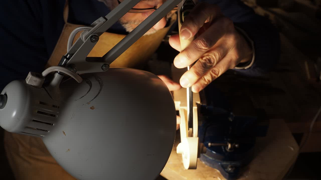 Luthier working intently under a bright lamp, illuminating the fine details of a violin scroll, held securely in a vise, using small scraper or chisel—to carefully refine inner curves and contours