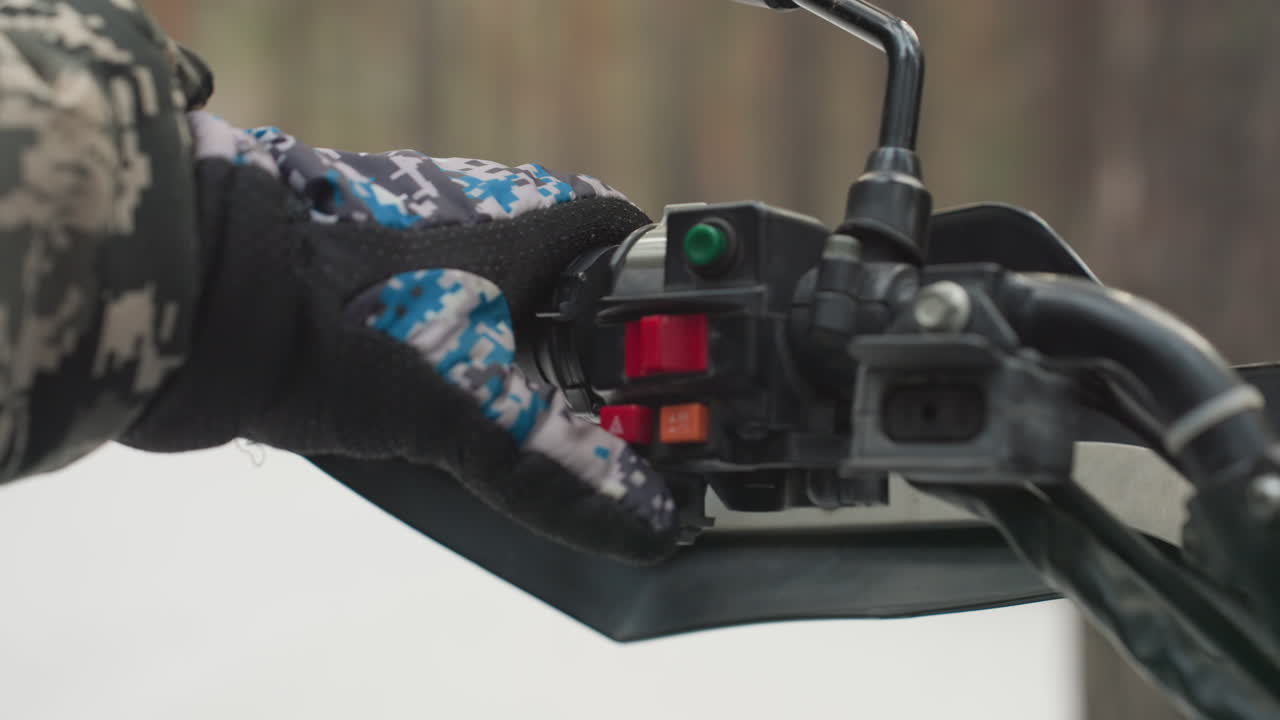 Mão enluvada no acelerador do ATV em fundo de floresta nevada. Piloto com jaqueta camuflada segura o guidão, dedo posicionado sobre o interruptor vermelho e indicador verde, espelho e retrovisor visíveis, close-up captura a luva.