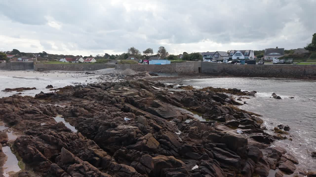 Low forward flight over rocky outcrops on shoreline of sandy beach with seabirds taking flight heading towards land on bright but cloudy day with interesting sky