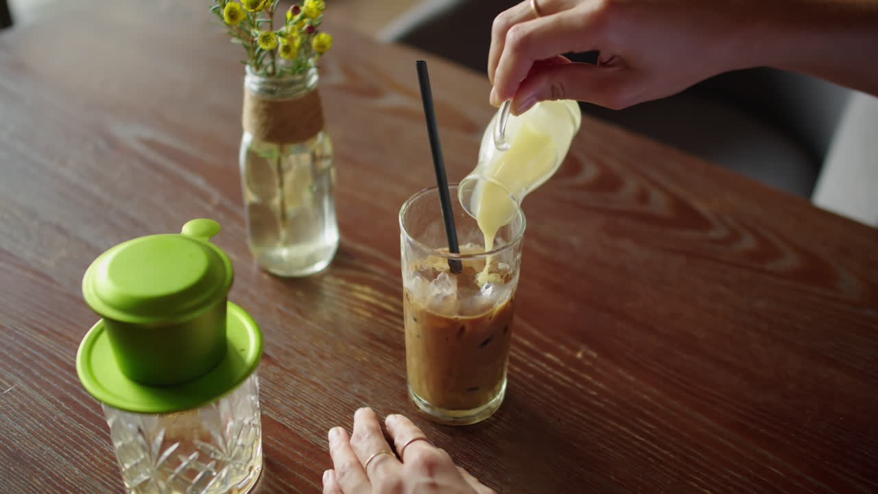 Person Adding Condensed Milk to Iced Coffee in Cafe