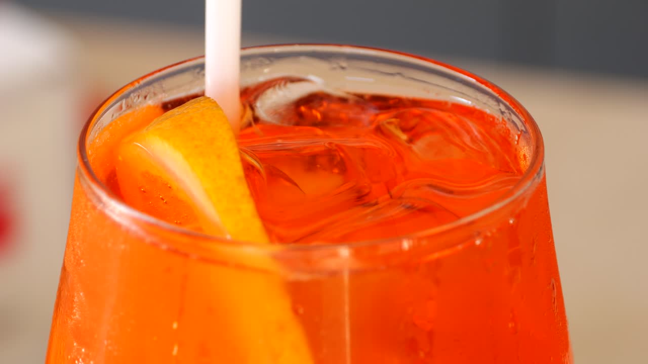 Primo piano di un vivace Aperol Spritz veneziano arancione con ghiaccio e una fetta di agrume mescolato con una cannuccia, ripreso in uno scatto dettagliato di una scena da bar