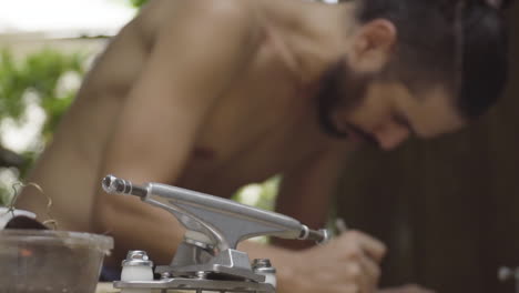 close-up in horizontal pan of skateboard truck with a bare-backed man drawing in the background