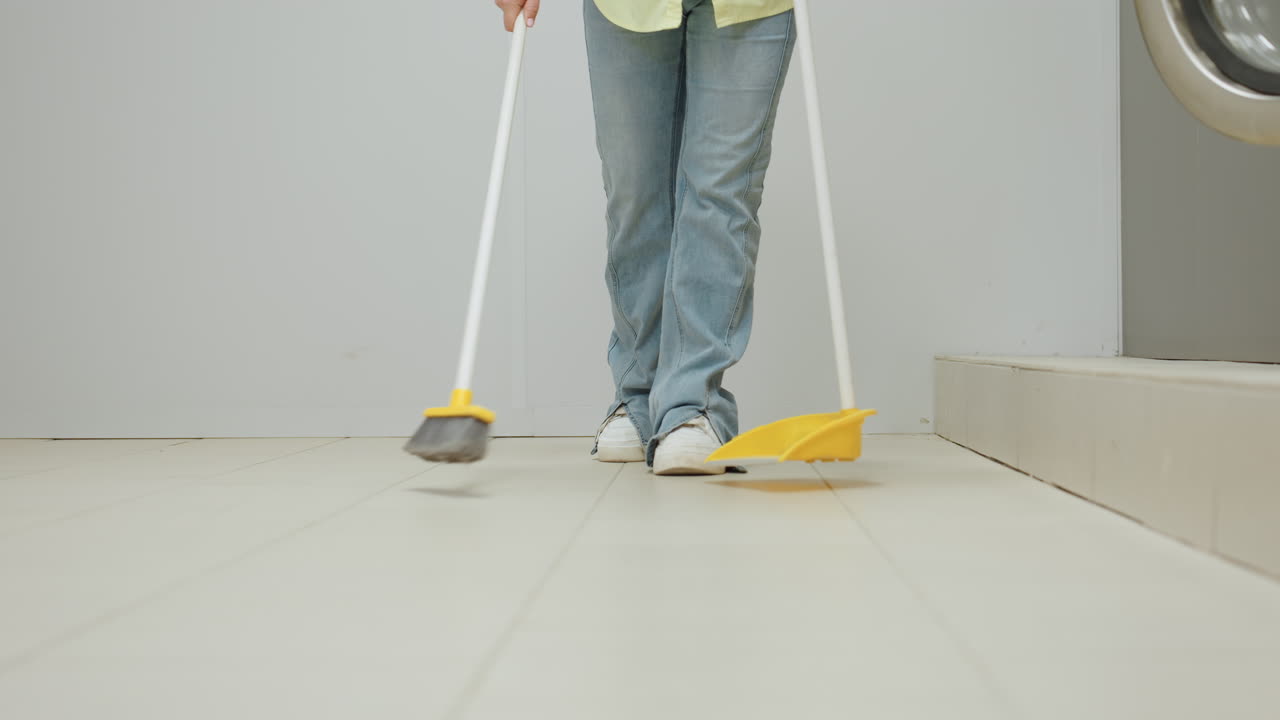 Long shot shows laundry owner in yellow shirt, jeans, white sneakers, sweeping tiled floor with broom and dustpan beside industrial washing machines, routine cleaning in bright interior