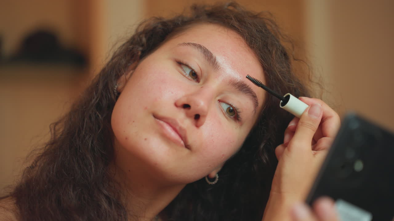 Casual face grooming in warm hotel lighting. Closeup of woman applying mascara in relaxed hotel environment. Woman adjusting her eyebrows using mascara in softly lit hotel room setting