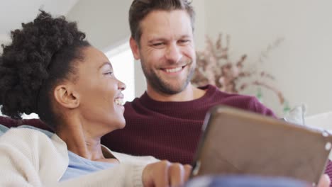 video de una feliz pareja diversa abrazando y usando una tableta en el sofá