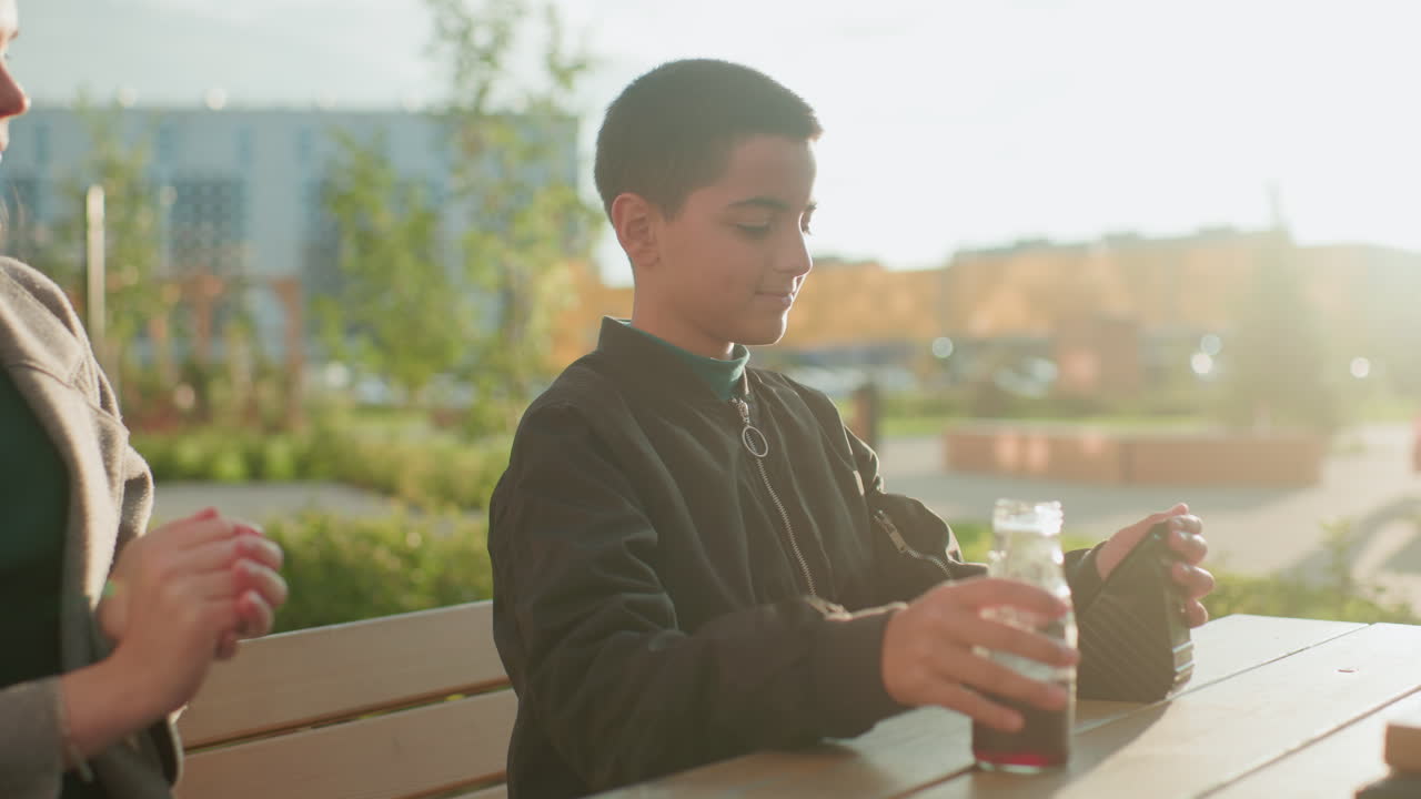 Kid collects lunch from elder sister outdoors, holding drink with warm expression of gratitude and joy, natural light glowing in background highlighting caring sibling bond