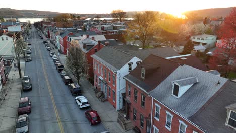 Slow-aerial-footage-of-a-neighborhood-in-a-small-town-in-America-at-sunset-with-the-sun-gleaming-through-the-trees-and-reflections-off-of-the-homes-and-buildings-ending-at-a-riverfront