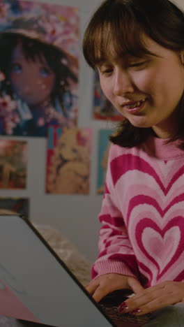 Vertical-view-of-teen-girl-in-headphones-typing-on-keyboard-and-chatting-with-friend-online-on-laptop-sitting-in-her-cozy-bedroom.-African-American-girl-spending-leisure-time-and-having-fun-at-home.