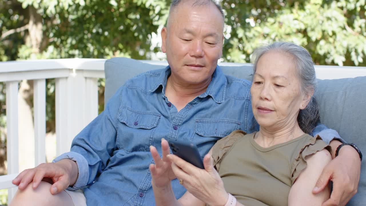 ベランダに座って、男性が携帯電話を見せ、女性がタップしてスクロールし、共有する年配のアジア人夫婦