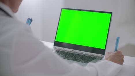 Eine-Ärztin-Berät-Sich-Online-Und-Schaut-Sich-Das-Grüne-Display-Eines-Laptops-An,-Um-Herauszufinden,-Ob-Sie-Chroma-Key-Technologie-Verwendet-Und-Notizen-In-Ein-Heft-Schreibt