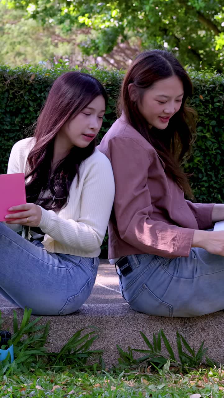 นักเรียนหญิงสองคนนั่งหันหลังชนกันในสวนสาธารณะ กำลังอ่านหนังสือและใช้แล็ปท็อป