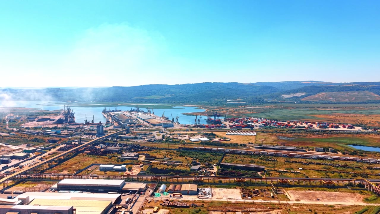 Oversiktsbilde av industrihavn og kystnært lagringskompleks. Lastcontainere og havnekraner står langs vannet under en klar blå himmel på dagtid.