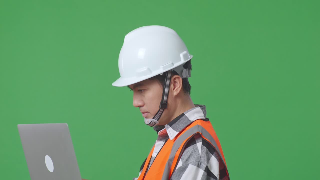 Close Up Side View Of Asian Male Engineer With Safety Helmet Working On A Laptop While Standing In The Green Screen Background Studio