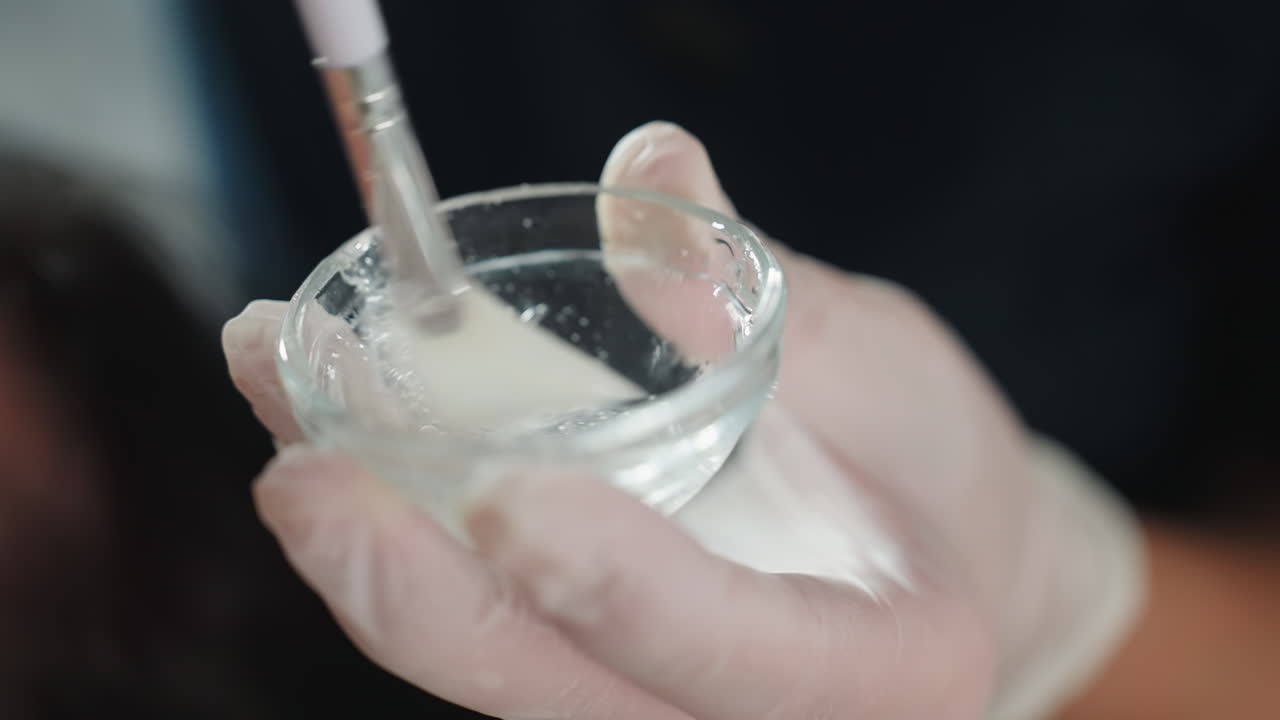Close up of gloved hands stirring clear gel with brush inside small glass bowl under bright spa light, showcasing bubble texture and professional skincare preparation for facial treatment session