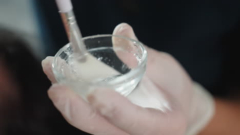 close up of gloved hands stirring clear gel with brush inside small glass bowl under bright spa light, showcasing bubble texture and professional skincare preparation for facial treatment session