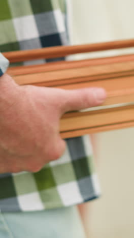 un primer plano de un hombre con una camisa a cuadros verde y azul sosteniendo un conjunto de listones de madera, revelando el grano de madera y los tornillos de oro. fondo borroso de una pared blanca y césped verde