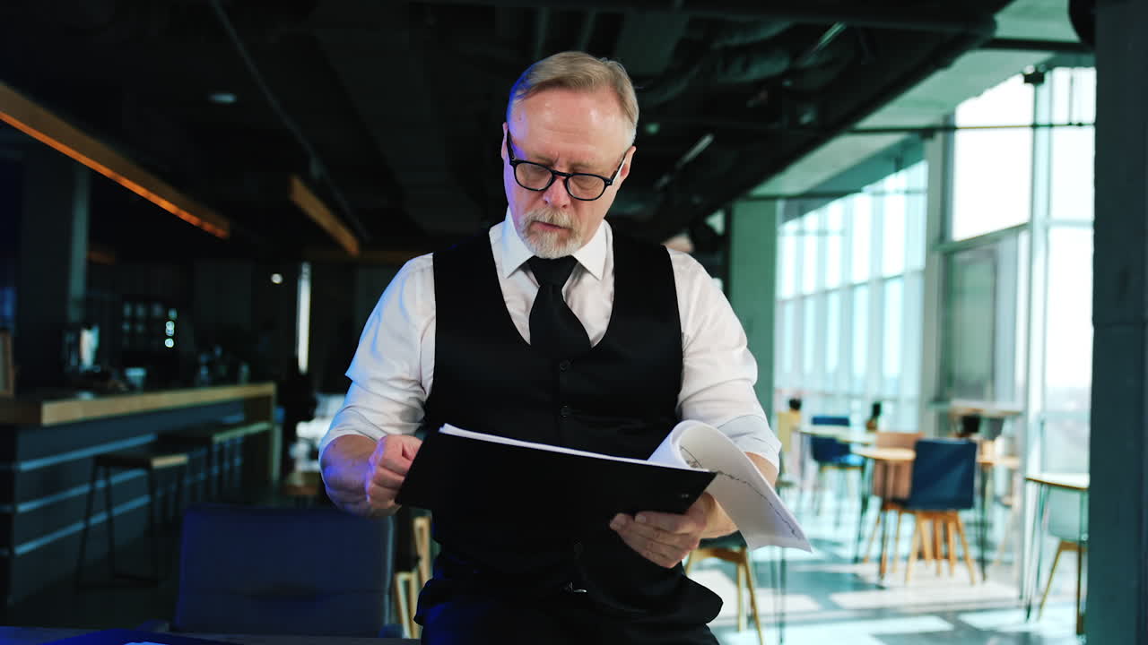 Serious focused businessman in white shirt and black vest holding papers. Bearded man turning the pages checking the information.