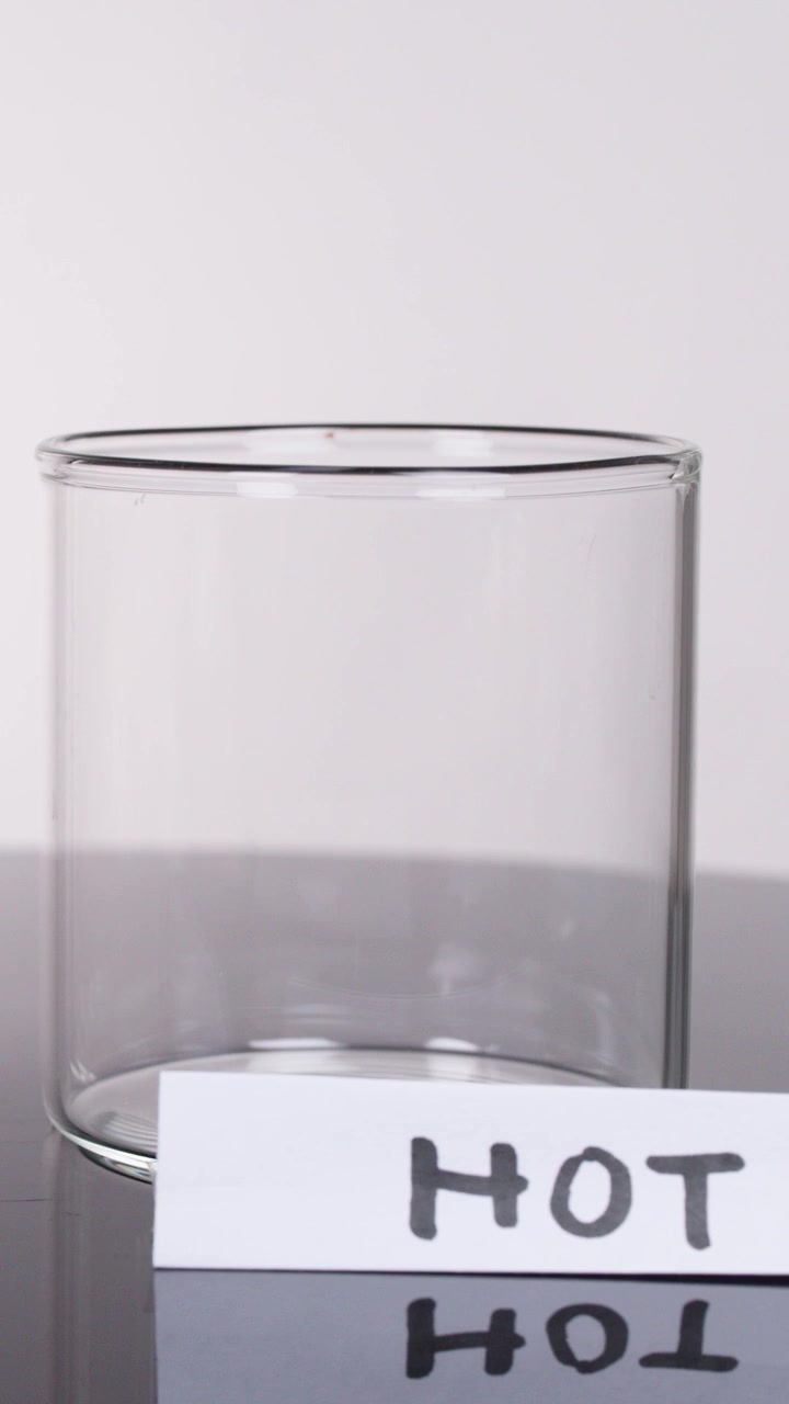 Una mano posiziona un foglio etichettato 'HOT' accanto a un becher di vetro vuoto su una superficie riflettente da laboratorio, inquadratura statica.