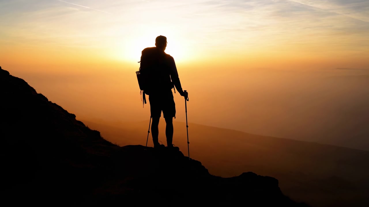 Un escursionista solitario sta con i bastoncini da trekking su una cresta aspra mentre la calda luce dell'alba risplende dietro la figura, creando una drammatica scena montana in controluce.