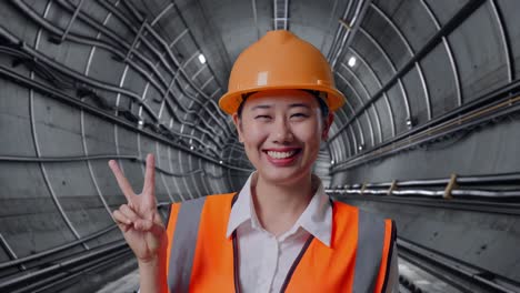 close up of asian female engineer with safety helmet smiling and showing peace gesture in underground subway tunnel