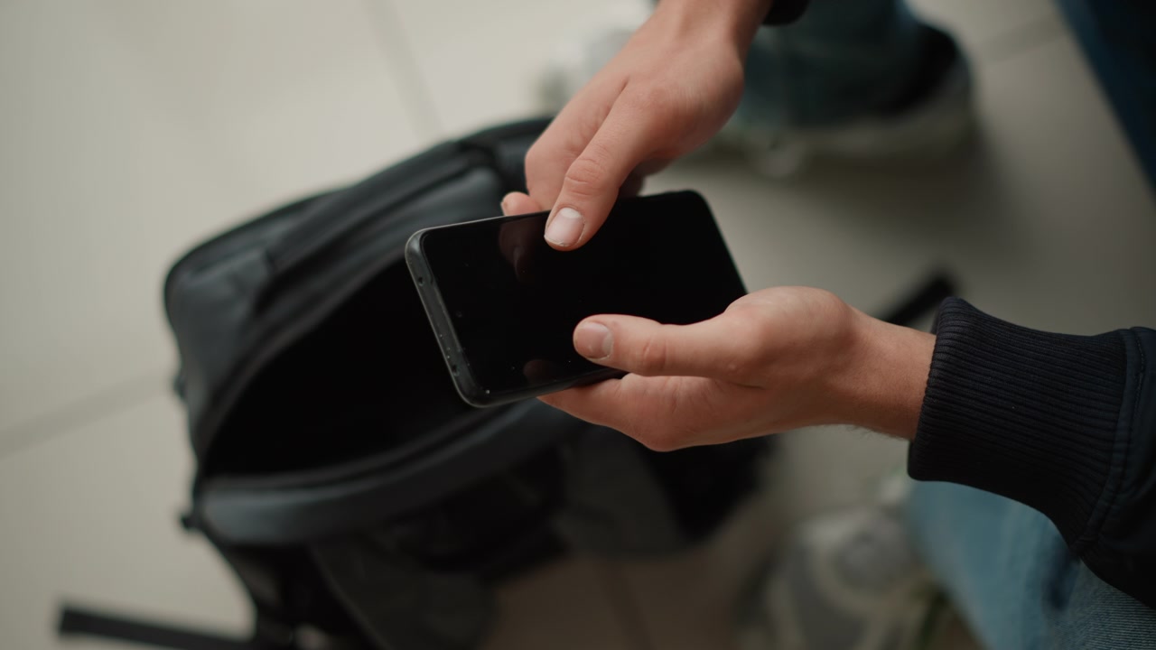 Close-up seseorang memegang smartphone di atas ransel hitam terbuka di lantai berwarna terang. Orang yang mengenakan pakaian gelap itu mengoperasikan perangkat sambil duduk atau berdiri.