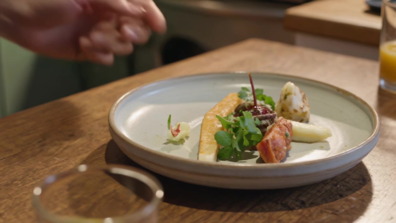 Chef con mangas verdes terminando de colocar salsa con una cuchara para emplatar en el mostrador, junto a pescado y verduras.