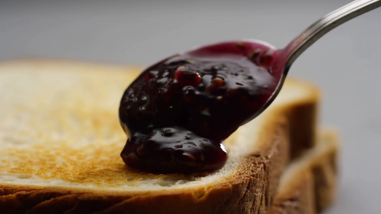 Close view captures spoon moving toward toast as dark fruit topping is placed on crisp bread, highlighting rich texture, warm colors, layered surface details, and simple kitchen food composition.