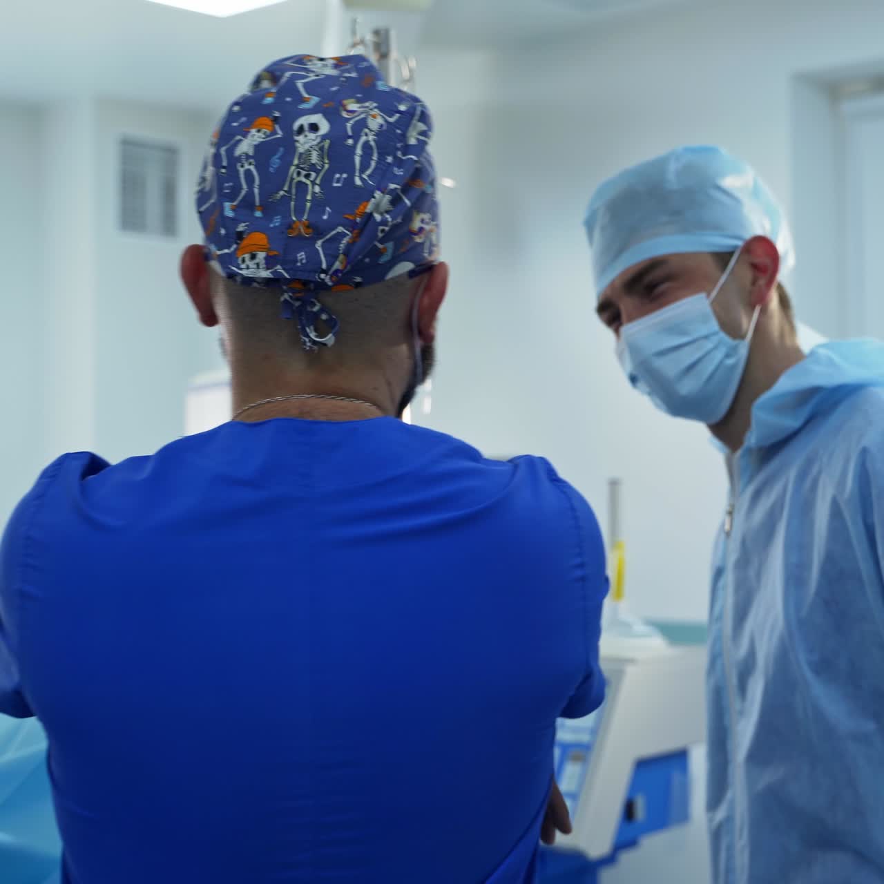 Two male doctors wearing different medical clothes stand in the surgery room. Surgery specialists stand their backs to the camera talking