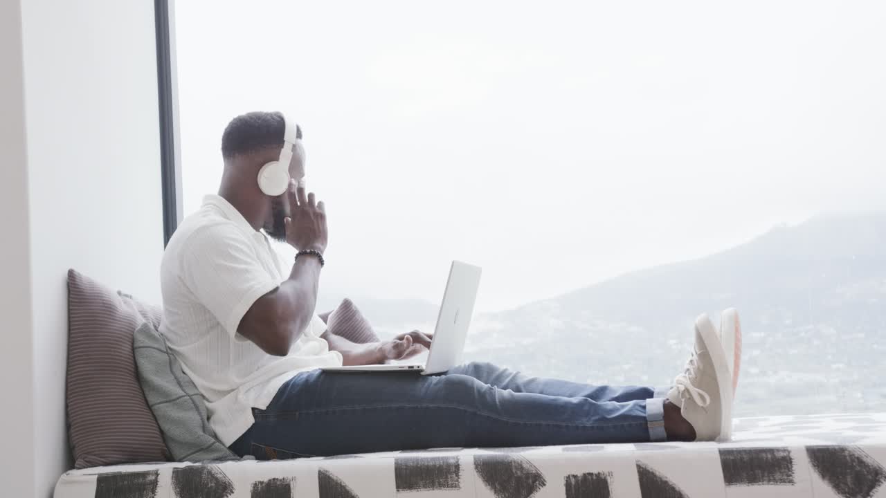 African American adult male typing on laptop at window bench, hearing cue, adjusting headphone