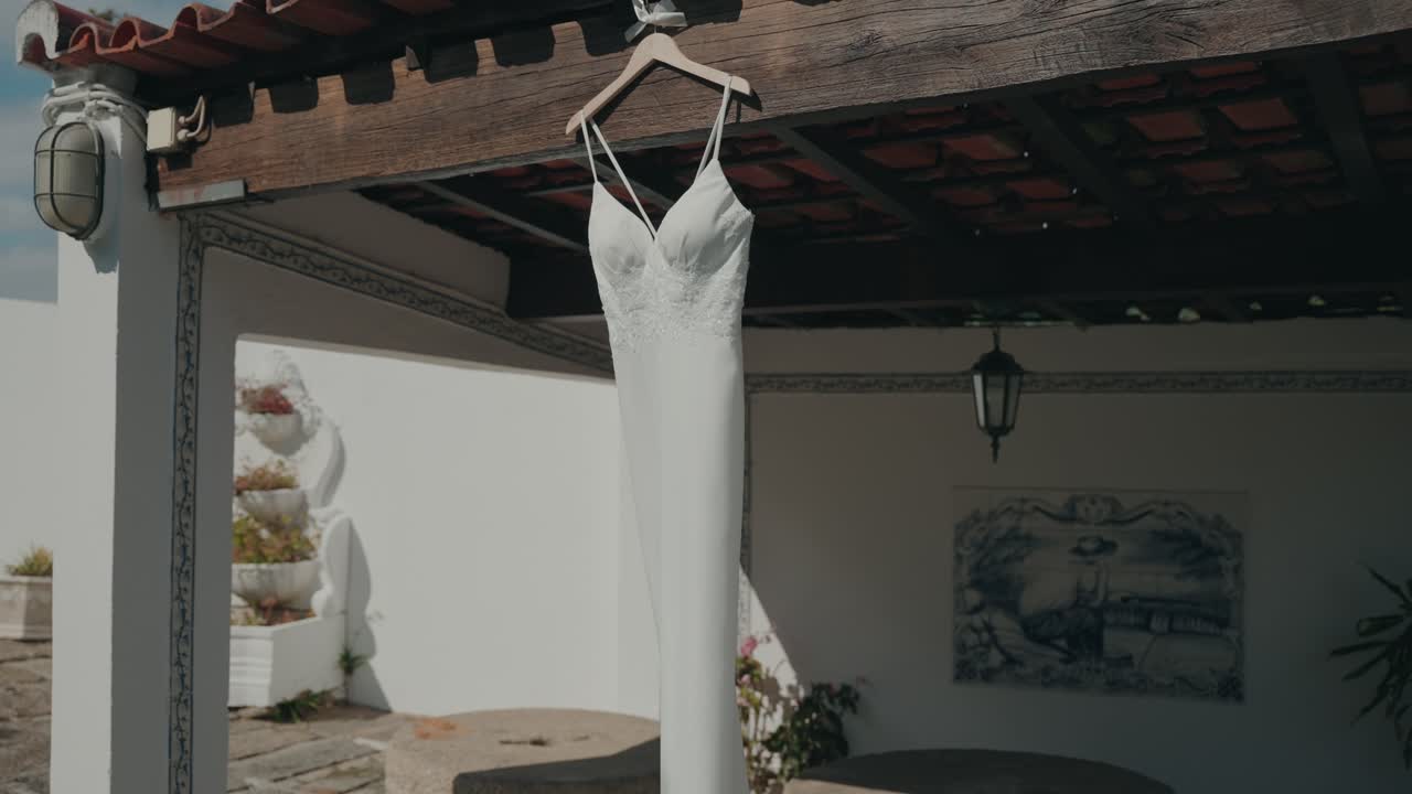 white wedding dress hangs from a wooden beam in a bright outdoor courtyard, softly lit by natural sunlight