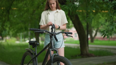 hermosa niña ajusta guantes negros al lado de su bicicleta en un camino pavimentado, rodeado de exuberante vegetación y árboles, con un fondo borroso de edificios y hojas revoloteando en la brisa