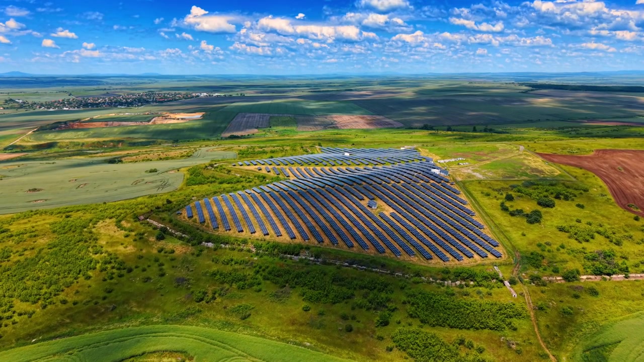 Buede rækker af solpaneler på en grøn bakke. Luftfoto panorama af en solcellepark omgivet af landbrugsmarker under en skyet blå himmel.