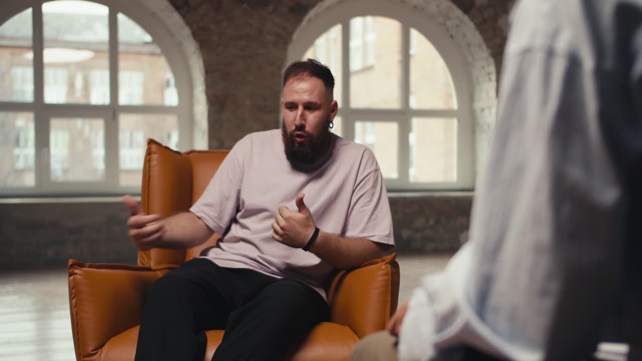 An excited man with a beard in a pink shirt talks about his problems and questions to a psychologist. A young man sits on a brown chair and communicates with a psychologist in a special room made of bricks