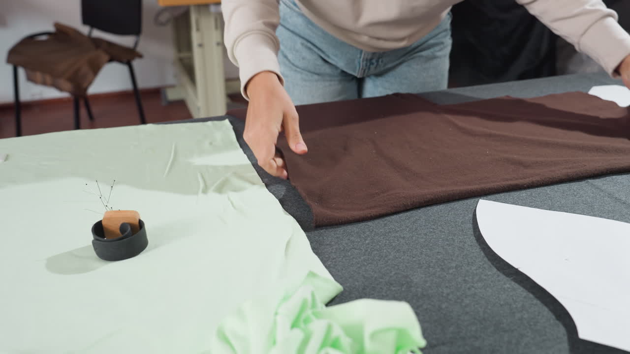 Female creative takes out light green fabric on work table while placing brown fabric nearby during garment preparation process in organized sewing studio with materials tools and bright environment