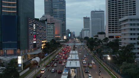 Low-flying-aerial-view-of-busy-multi-lane-road-with-car-traffic-near-roundabout-in-the-evening-with-beautiful-city-lights-glowing-in-downtown-Jakarta,-Indonesia