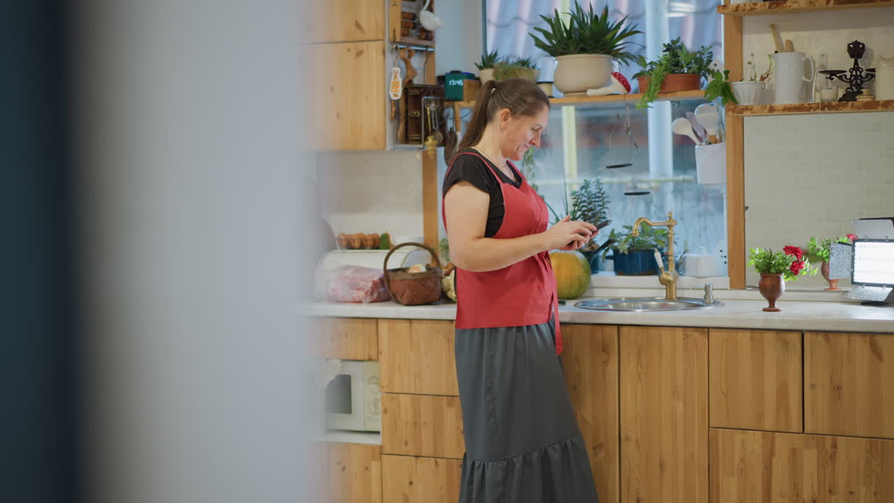 Scatto spontaneo dalla porta di un cuoco casalingo che si prepara, attraverso la porta sfocata una donna controlla il telefono e misura gli ingredienti su un bancone di legno, piante e gadget da cucina su uno scaffale, morbida retroilluminazione dalla finestra, momento quotidiano.